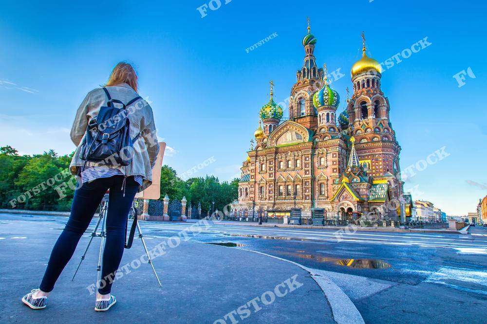 На фотографии изображён художник, стоящий перед храмом Спаса-на-Крови в Санкт-Петербурге. Раннее утреннее солнце освещает яркие купола храма, подчёркивая его богатое убранство и уникальную архитектуру. Художник с мольбертом передаёт дух творчества и вдохновения, который царит в этом историческом месте. На фоне видны деревья и чистое голубое небо, добавляющие гармонии городской сцене.